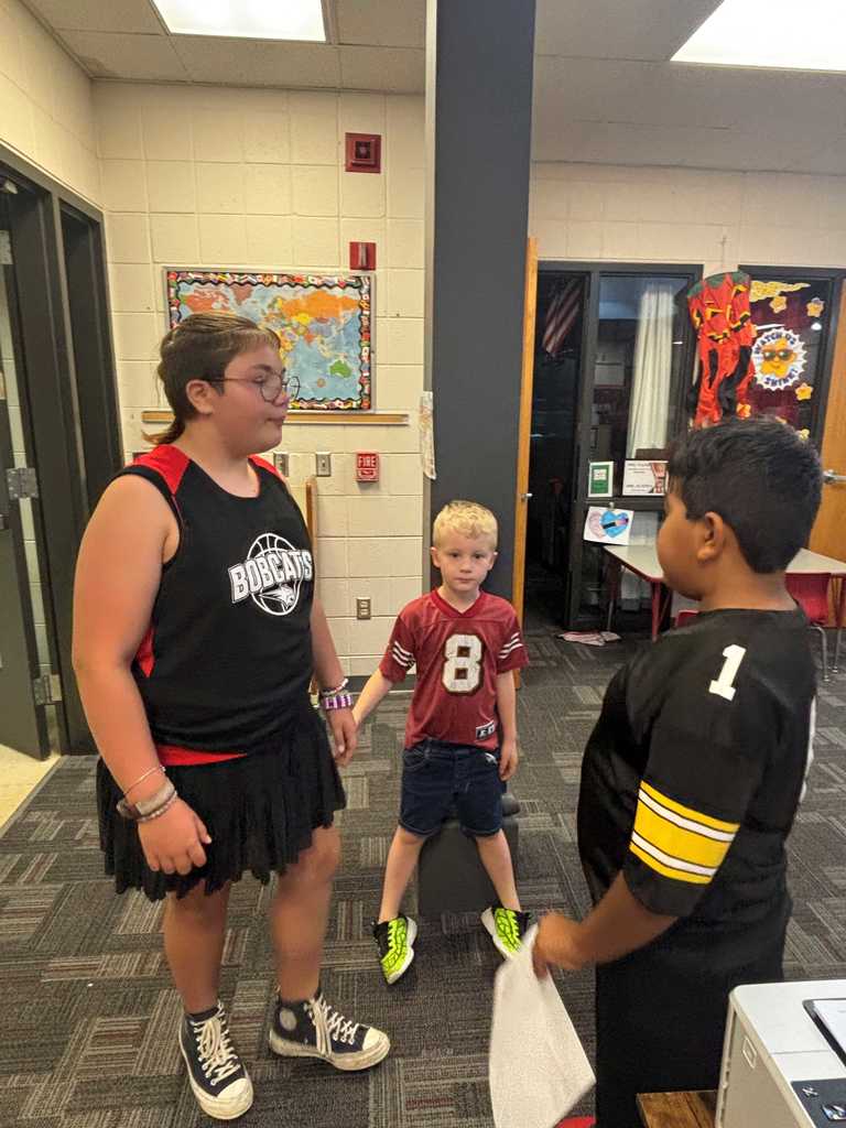 Three children stand in a classroom, wearing sports jerseys with numbers.