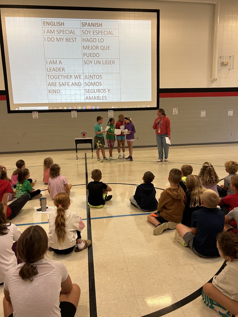A group of students presents in front of peers seated on a gym floor. A large screen shows English and Spanish affirmations: "I am special."