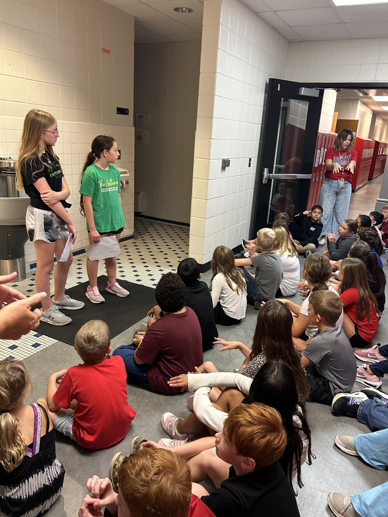 Two young girls stand at the front of a classroom, speaking to seated children.
