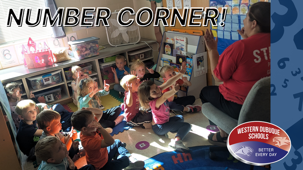 Children sit on the floor in a classroom, attentively watching a teacher in red, holding up numbers.