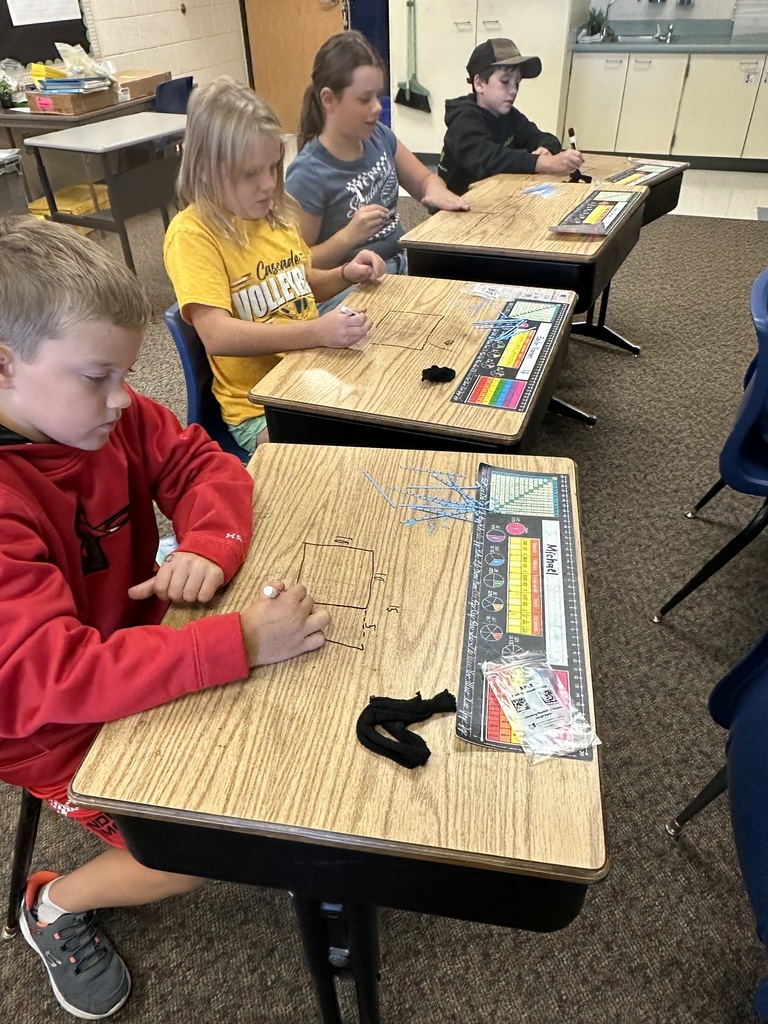 Four children sit at desks in a classroom, using whiteboard markers to write on desk surfaces.