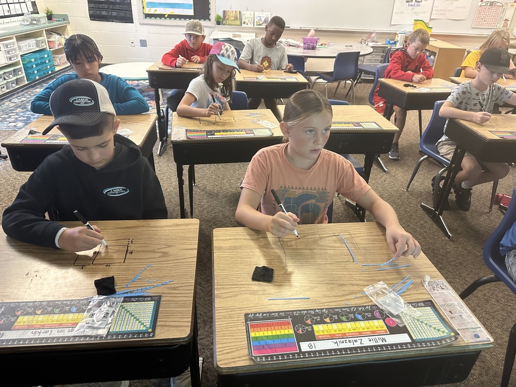 Classroom scene with students seated at desks drawing with thin blue sticks and markers.