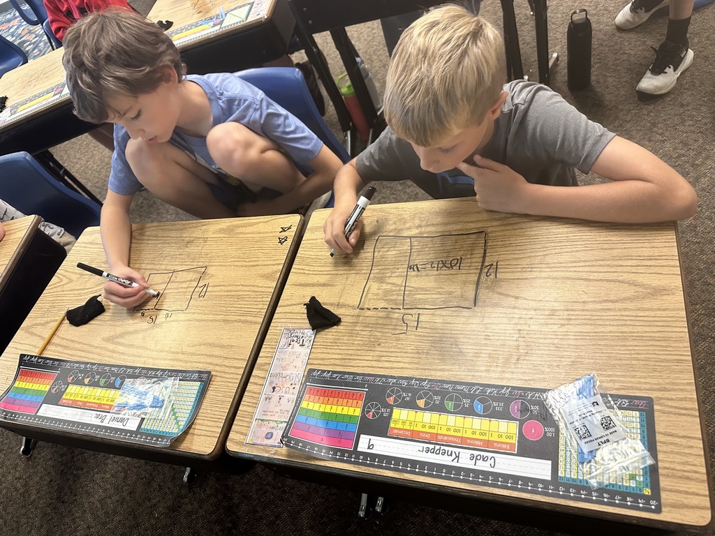 Two boys in a classroom draw geometric shapes on desks with markers.