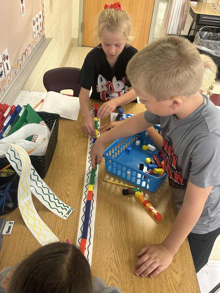 A group of elementary students stand around a classroom table, using colorful plastic cubes to measure a long paper strip marked with a wavy line pattern.