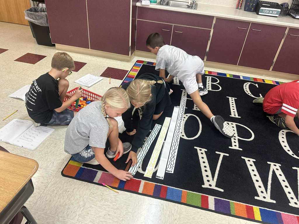 Several students sit and kneel on a classroom rug with alphabet letters, using plastic cubes and paper strips with wavy lines as they work on a math activity together.