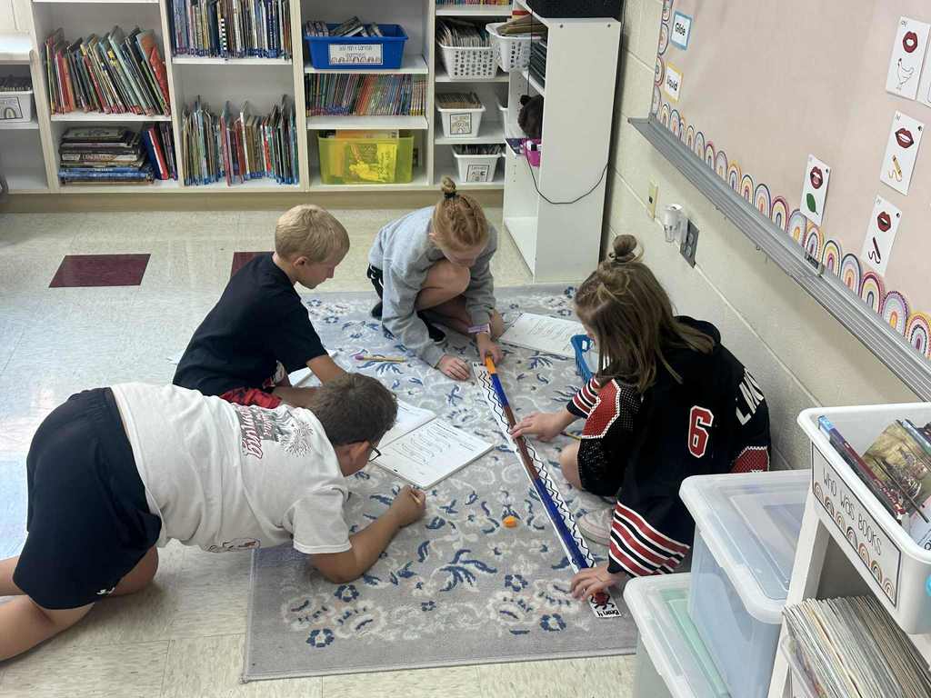 Four students sit and kneel on a light-colored classroom rug near a bookshelf, aligning colorful cubes along a long paper strip while writing on worksheets.