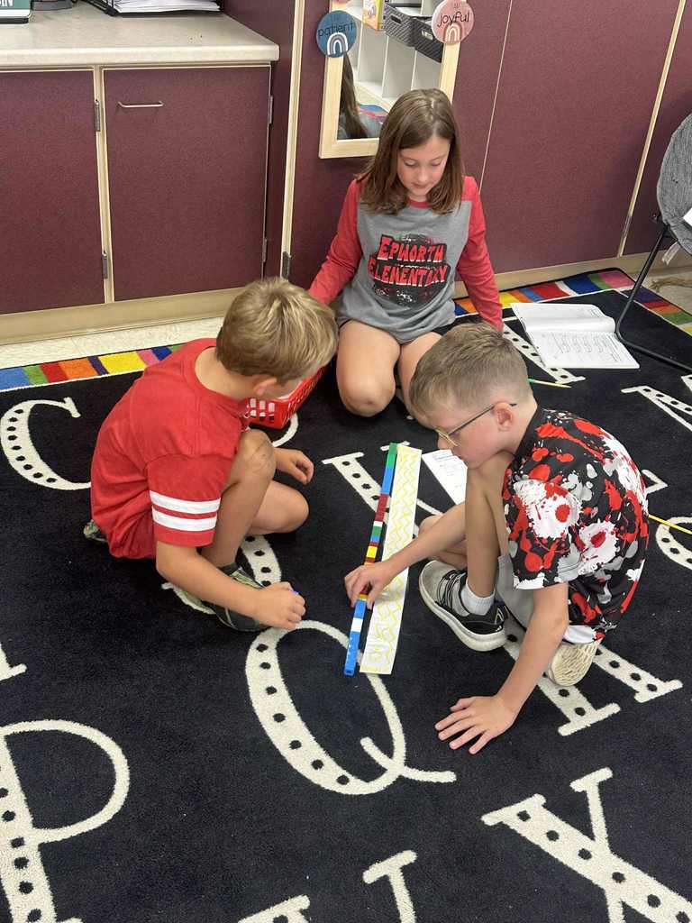 Three students sit on the floor of a classroom alphabet rug, placing blocks along a paper strip with a yellow wavy line pattern as part of a group math activity.