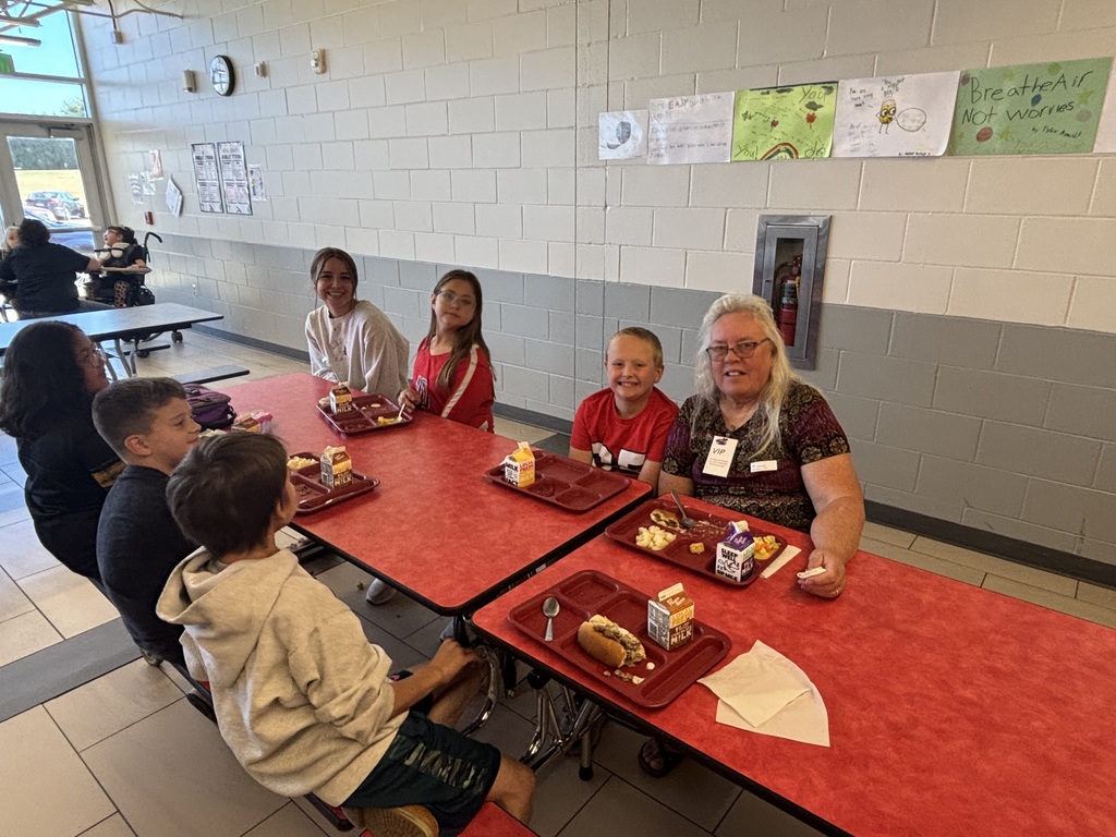 A group of students and two adults sit together at a cafeteria table with lunch trays, smiling at the camera with student artwork displayed on the wall behind them.