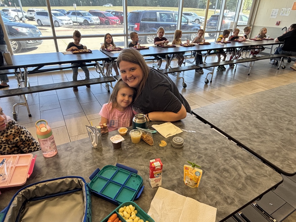 A young child and an adult sit close together at a lunch table, smiling while eating.