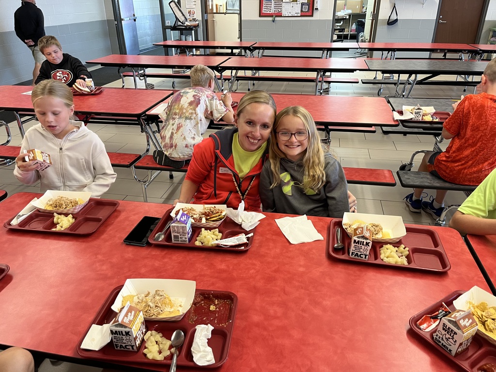 A student and an adult sit side by side at a red lunch table in a school cafeteria, smiling at the camera while eating lunch with other students seated around them.