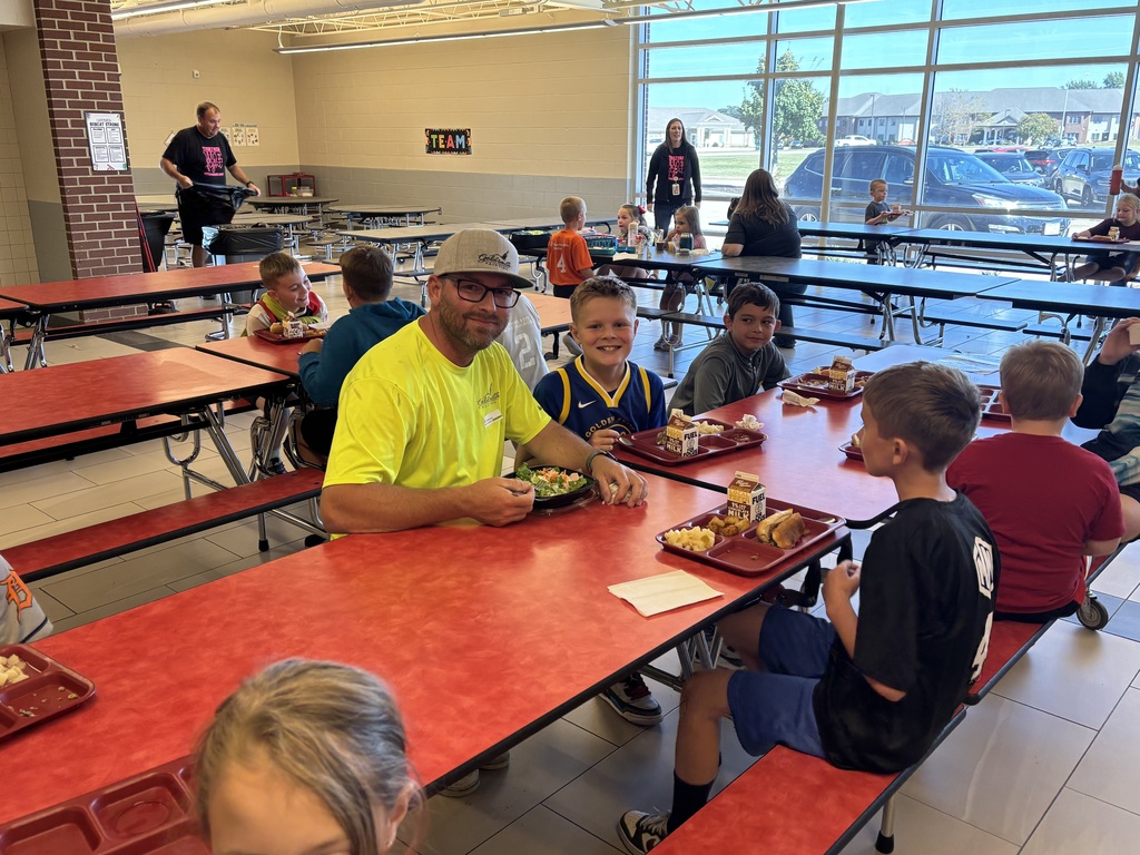 An adult wearing a yellow shirt sits at a cafeteria table eating with a group of students, all smiling and talking during lunchtime.