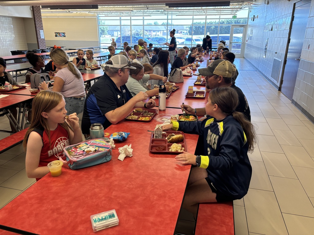 A cafeteria full of students and adults sits at long red lunch tables, enjoying their meals together during a family lunch event.