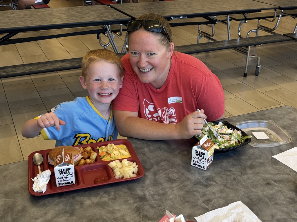 A young student grins widely next to an adult wearing a red shirt as they eat lunch together at a cafeteria table.