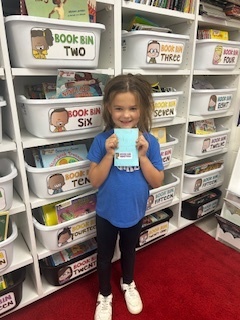 A smiling girl stands in front of organized classroom book bins, proudly holding up a small book she created.