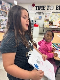 A young girl stands in a classroom holding an open notebook with drawings and writing, speaking while another student sits nearby watching.