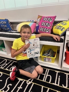 A boy seated on the classroom rug leans against a bookshelf, smiling and holding up a drawing or story he created.