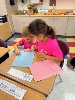 Two kids sit close together at a desk, looking at a handmade booklet as one points to the page while reading.