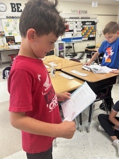 A boy in a red shirt stands next to a desk in a classroom, reading from a notebook while other students work around him.