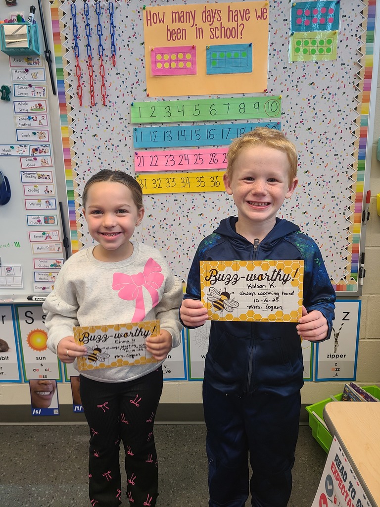 Two young students stand in front of a colorful classroom wall display showing numbers and a chart titled “How many days have we been in school?” They are smiling and holding “Buzz-worthy” award certificates.