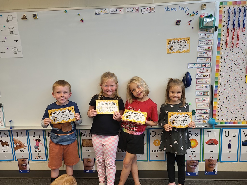 Four young students stand in a row in front of a whiteboard, smiling and holding “Buzz-worthy” award certificates. Classroom visuals and alphabet cards line the wall behind them.