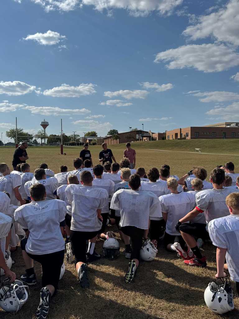 A large group of football players in white practice jerseys kneels on a grassy field facing a few older students or coaches who are standing and speaking to them.