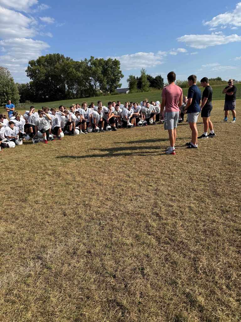 Football players wearing white jerseys sit or kneel in rows on the grass while three older students or coaches stand in front of them speaking.