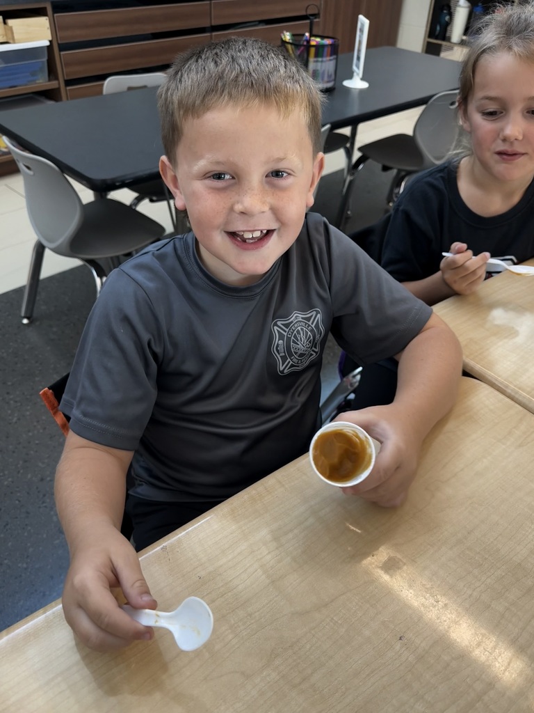A smiling young boy sits at a classroom table holding a small cup of caramel-colored pudding and a spoon, looking toward the camera.
