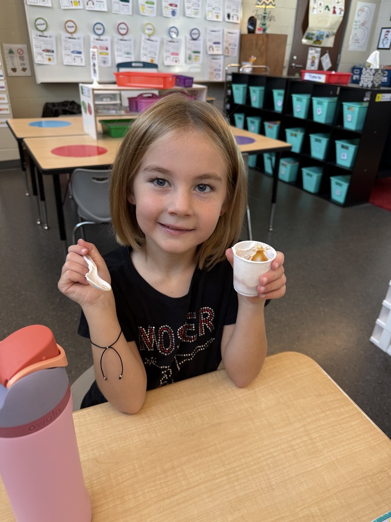 A young girl with a short blonde bob haircut sits at a classroom desk holding a small cup of pudding in one hand and a spoon in the other, smiling at the camera.