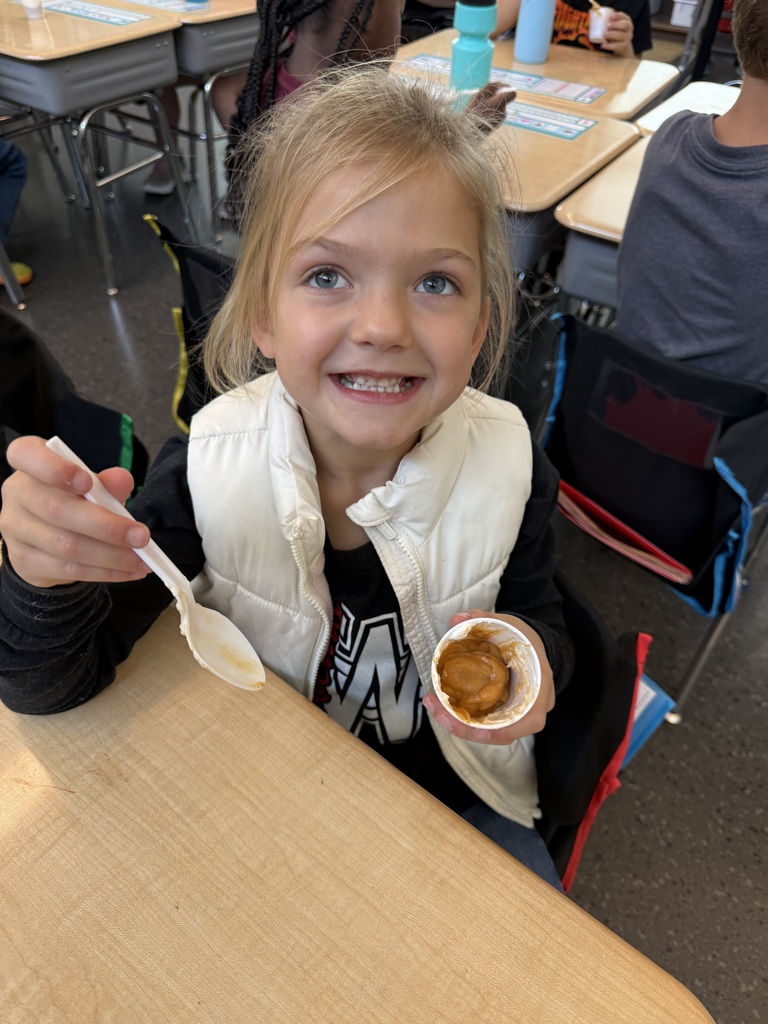 A smiling young girl wearing a white vest holds a cup of pudding and a spoon at her desk.