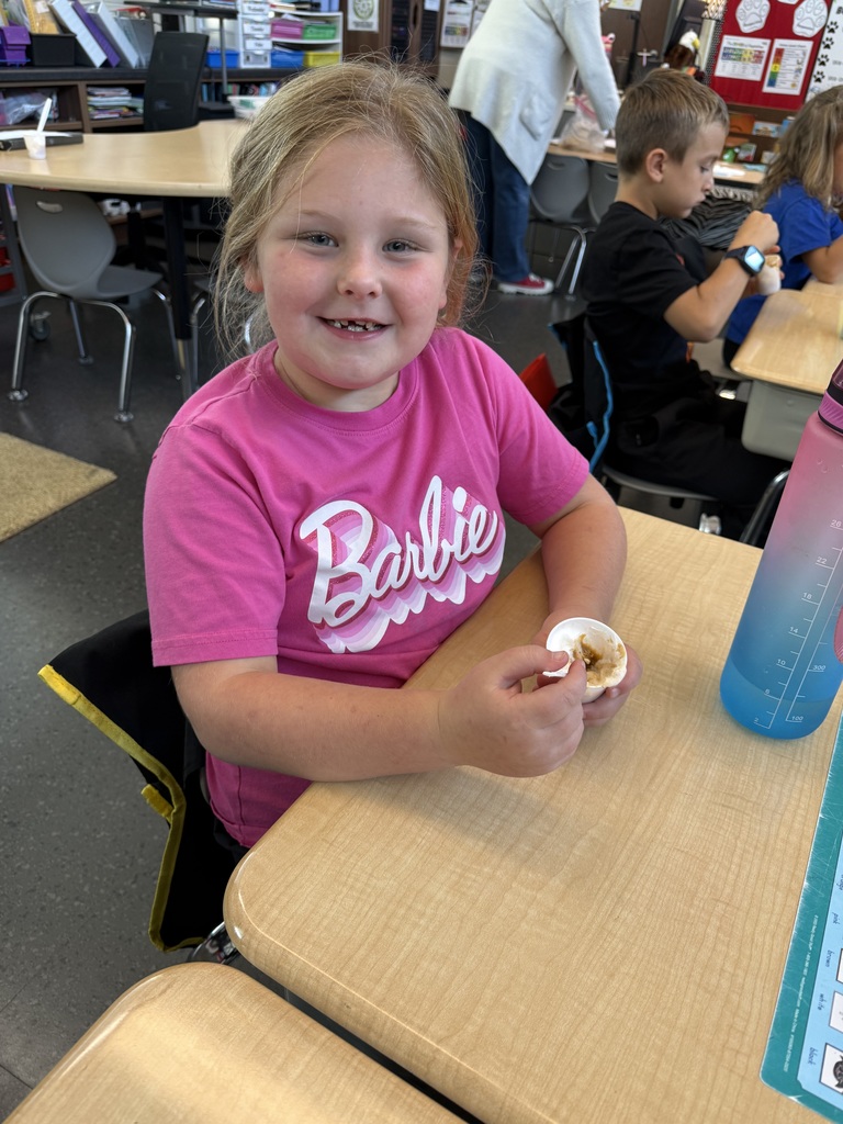 A smiling young girl wearing a pink Barbie shirt sits at her desk holding a small cup of pudding with a spoon.