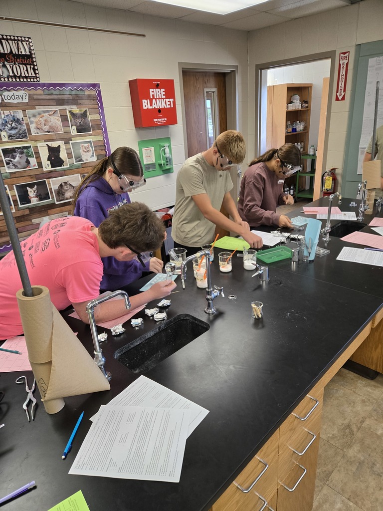 Four students wearing safety goggles work at a science lab counter, bending over worksheets and foil-wrapped samples.
