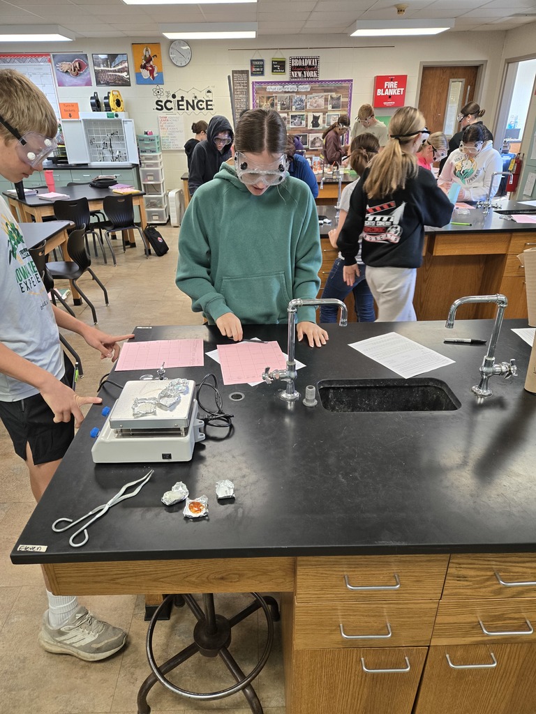 A group of students wearing safety goggles work at a science lab table.