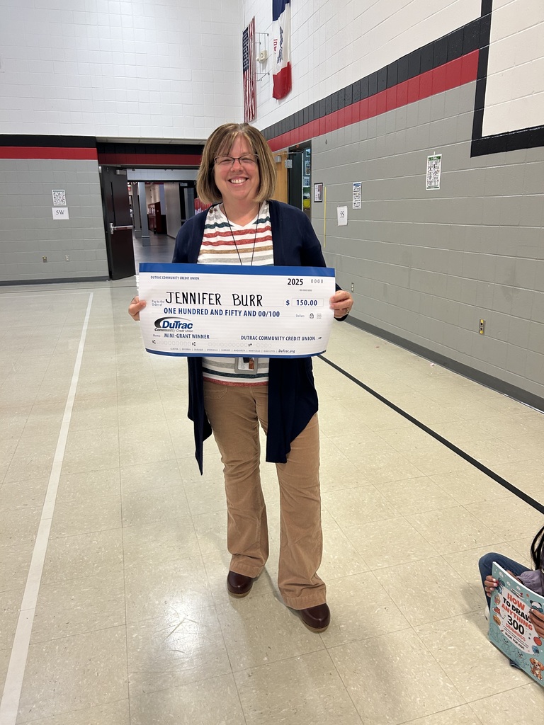 A smiling adult stands in a gymnasium holding an oversized check labeled “Jennifer Burr” for $150 from DuTrac Community Credit Union.