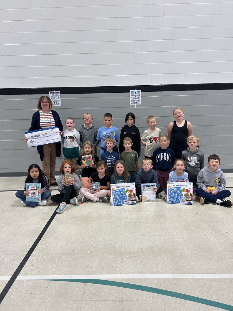 A group of elementary students sit and kneel in rows in a gymnasium holding learning materials and books. An adult stands on the left holding an oversized grant check. The students smile toward the camera.