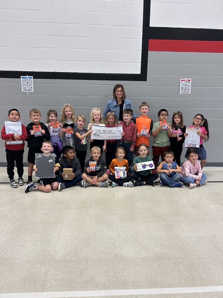 A group of young students sit and kneel in a gymnasium holding classroom supplies and learning items. An adult stands behind them in the center holding an oversized grant check labeled “Lori Messer.”