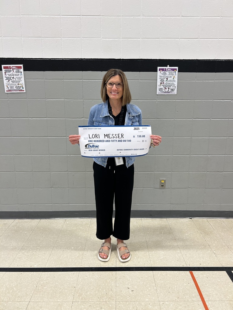 A smiling adult stands alone in a gymnasium holding an oversized check labeled “Lori Messer” for $150 from DuTrac Community Credit Union.