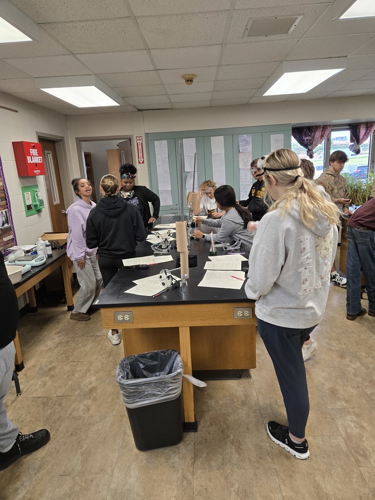 A group of high school students gather around a science lab table covered with lab papers and equipment.