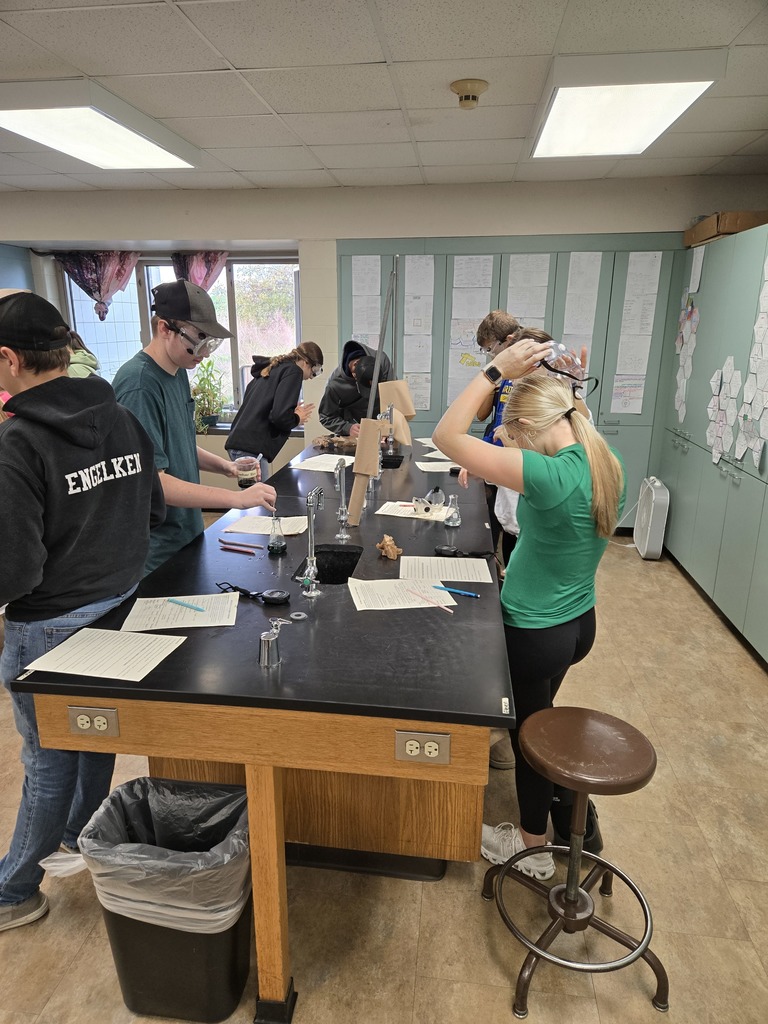 High school students wearing safety goggles work around a black science lab table with worksheets, beakers, and lab equipment.