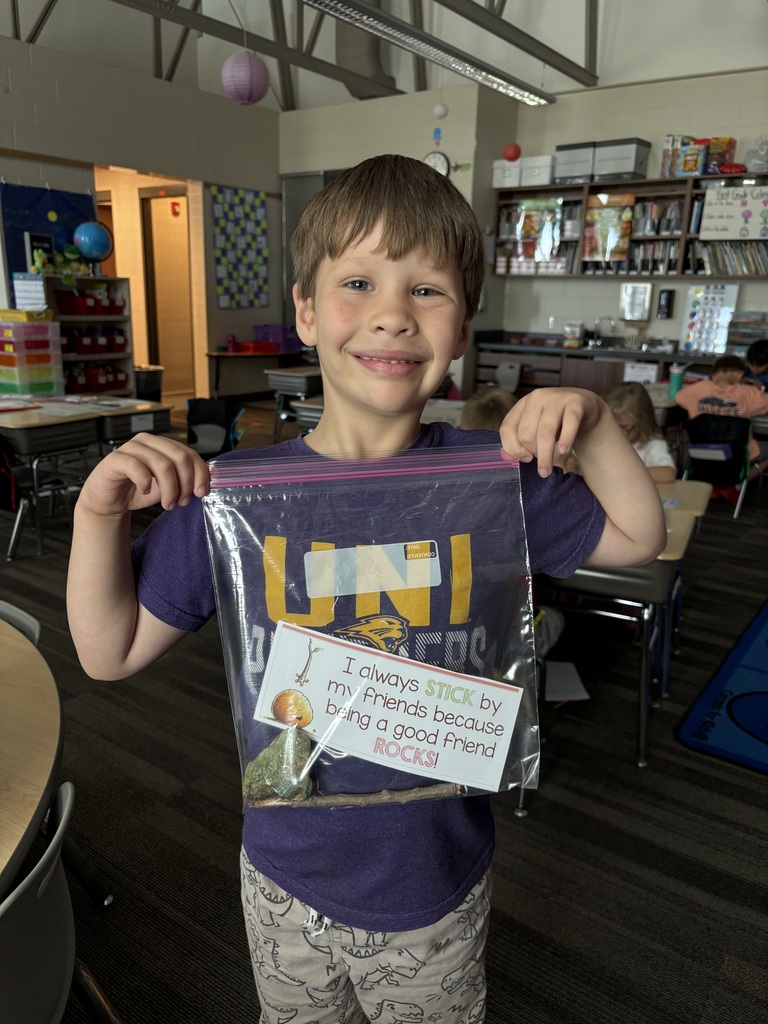 A smiling young student stands in a classroom holding up a clear plastic bag with a label that reads, “I always stick by my friends because being a good friend ROCKS!” The bag contains a stick, a painted rock, and a small craft.