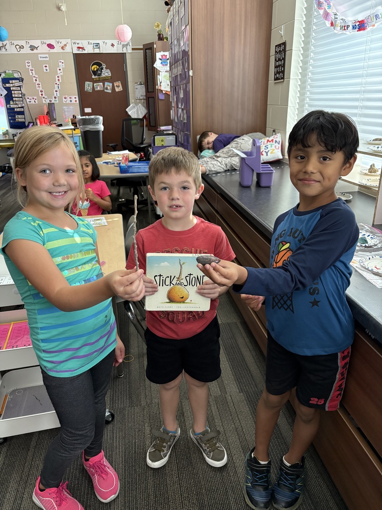 Three young students stand side by side in a classroom. The student in the middle holds the book Stick and Stone while the two students on either side hold up small rocks.
