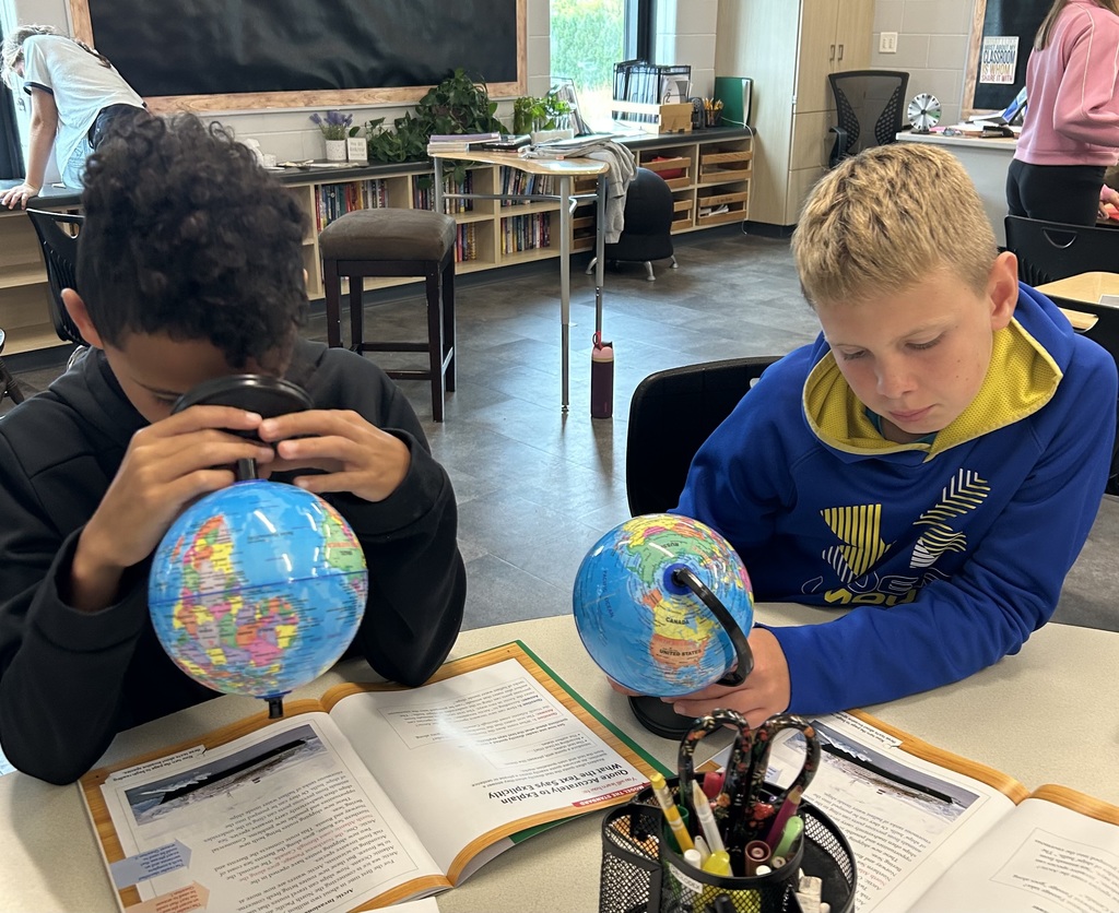 Two students sit at a classroom table with open textbooks and small globes. One student looks closely at the globe through a magnifying lens while the other studies the globe in his hands.