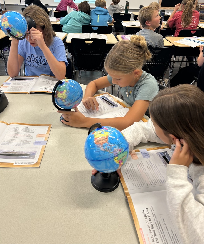 Three students sit together at a table with open textbooks and small classroom globes. One student uses a magnifying lens on the globe while the others read and point to the text.