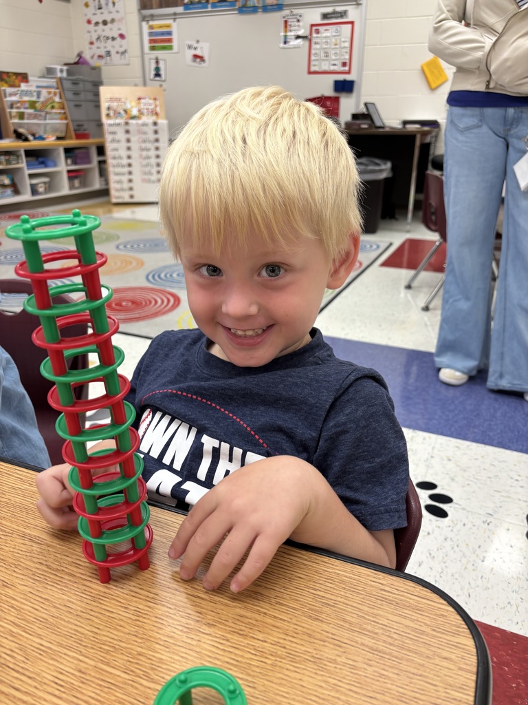 A smiling child sits at a classroom table and proudly holds a tall tower built from red and green interlocking plastic rings.