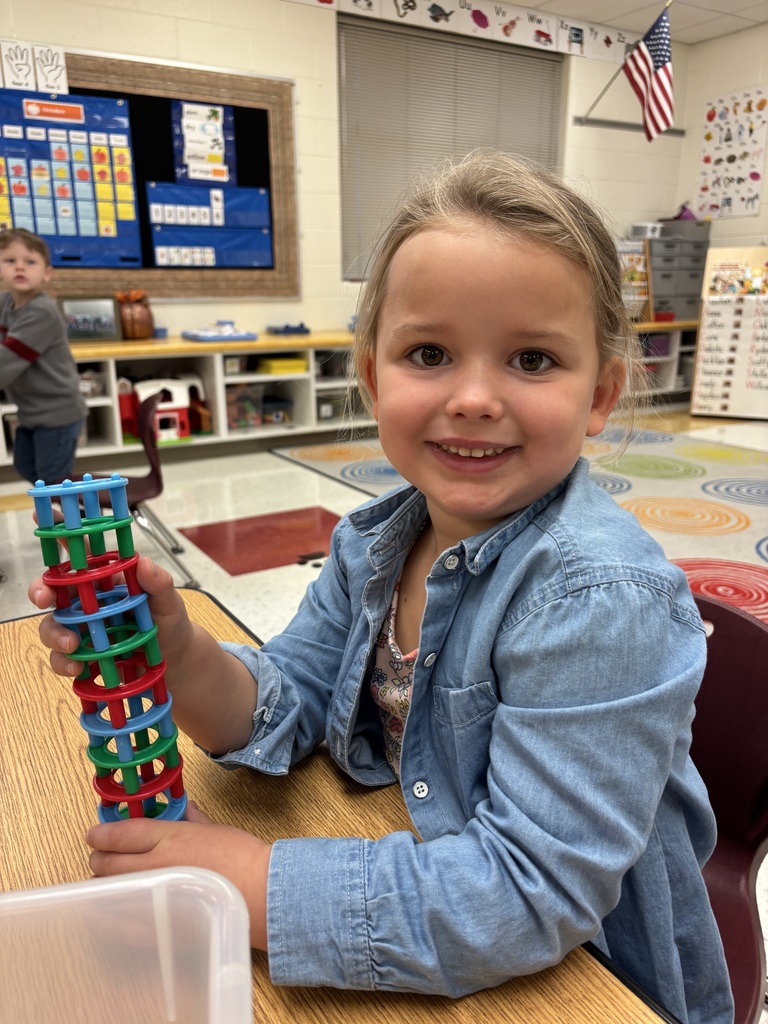 A smiling young girl sits at a classroom table holding a tall stack of blue, green, and red interlocking plastic rings she has built.
