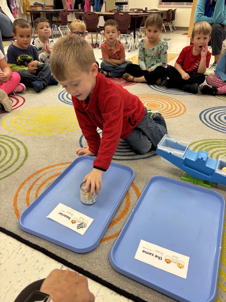 A young boy kneels on a colorful classroom rug and places a can on a blue tray labeled “heavier” while classmates sit in a circle behind him, watching the activity.