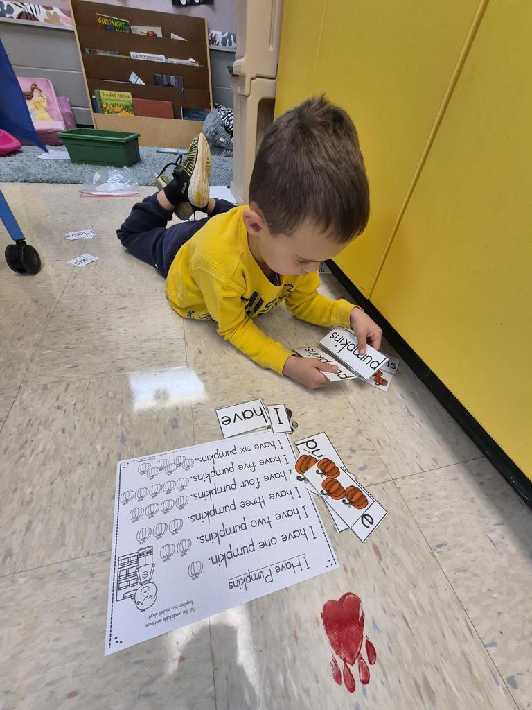 A student wearing a yellow shirt sits on the floor sorting and reading pumpkin word cards for a counting and reading activity.