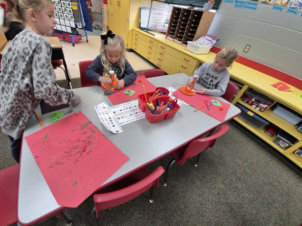 Three children sit at a classroom table working on an orange-and-black pumpkin craft using scissors, paper, and glue.