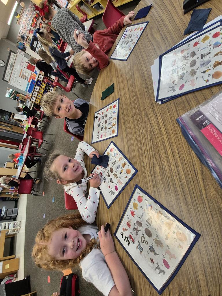 Four smiling students sit around a table working on alphabet recognition mats with dry-erase markers in a colorful classroom setting.