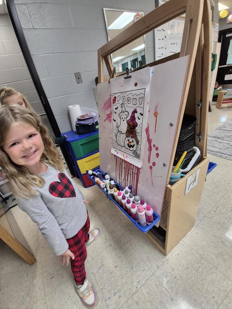 A smiling student stands beside an easel displaying her Halloween-themed painting featuring a ghost and a pumpkin.