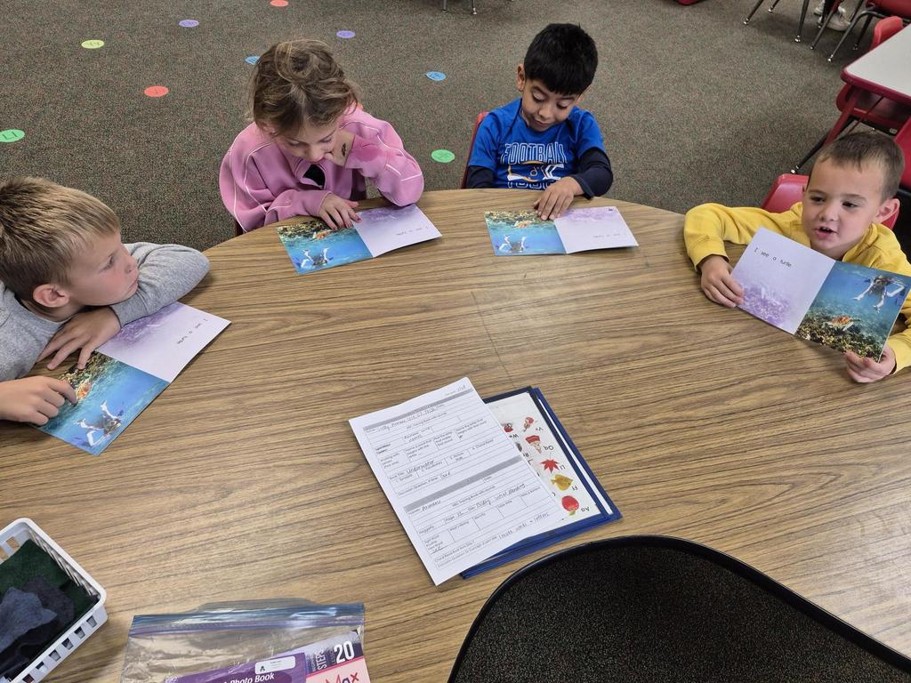 Four young students sit around a table reading illustrated books about the ocean. Worksheets and folders are spread out on the table.